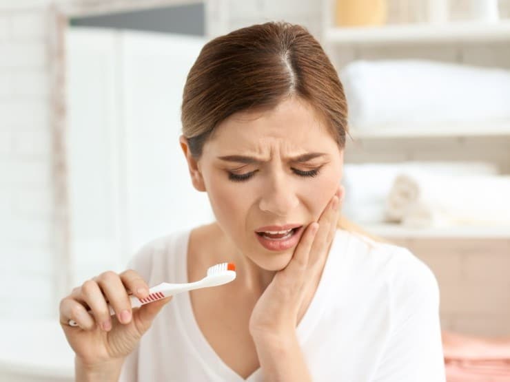 Woman holding toothbrush to cheek, experiencing tooth pain in bright dental office setting.