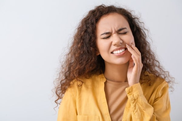 Woman in yellow shirt touching her cheek, appearing to experience tooth pain or discomfort.