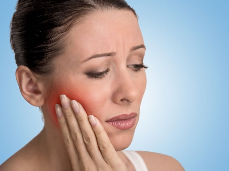 Woman with hand on cheek experiencing tooth pain or jaw discomfort against blue background.