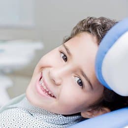 Young child smiling at dental office during examination with blue dental equipment visible.