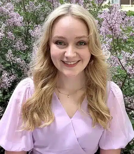 Young woman smiling at camera with purple flowers blooming behind her outdoors.