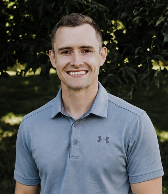 Dr. Daniel Clark smiling in a grey polo shirt with a leafy green background.